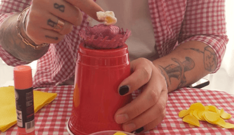 Pessoa decorando copo de plástico vermelho com mini chapéu de festa junina e pipoca