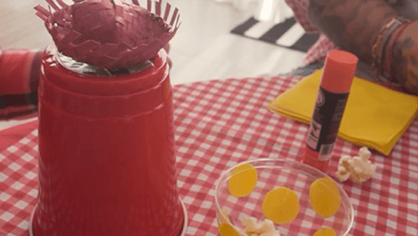 Mini chapéu caipira sobre copo de plástico vermelho, em mesa com toalha xadrez e itens de artesanato