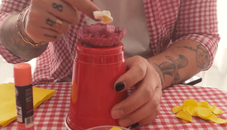 Decoração de copo vermelho com pequeno chapéu de palha roxo em uma mesa com itens de artesanato