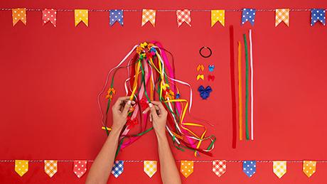 Mãos amarram fitas coloridas em um elástico de cabelo, preparando um enfeite de cabeça para festa junina, para ser utilizado como rabo de cavalo