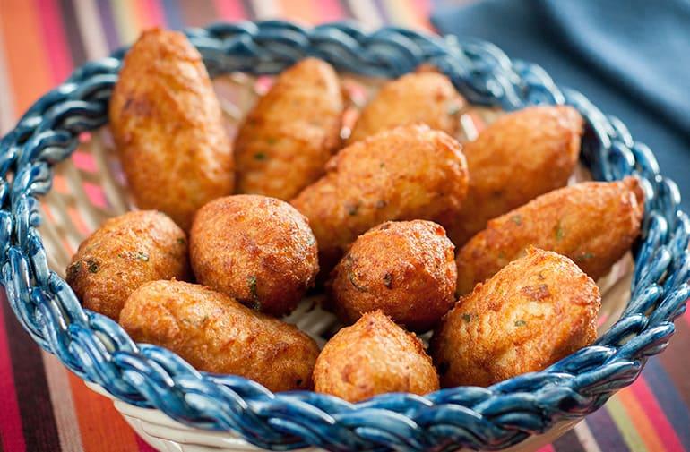 Porção de bolinhos de bacalhau fritos e dourados, servidos em cesto de madeira rústico com detalhes azuis. Petisco português