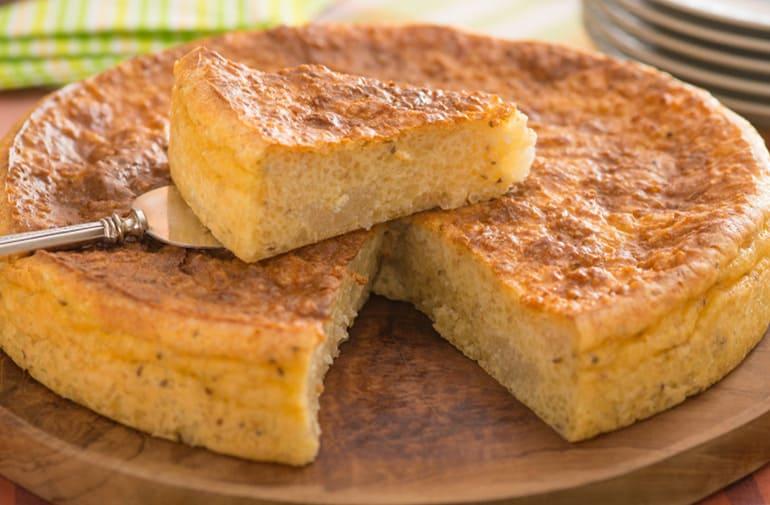 Bolo de tapioca cremoso, massa fofinha, servido sobre tábua de madeira, com uma fatia sendo retirada