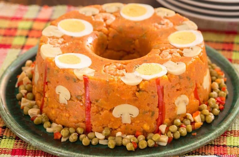 Cuscuz paulista no formato de pudim, servido sobre um prato verde e decorado com ovos cozidos, ervilha, tomate, pimentão e champignon