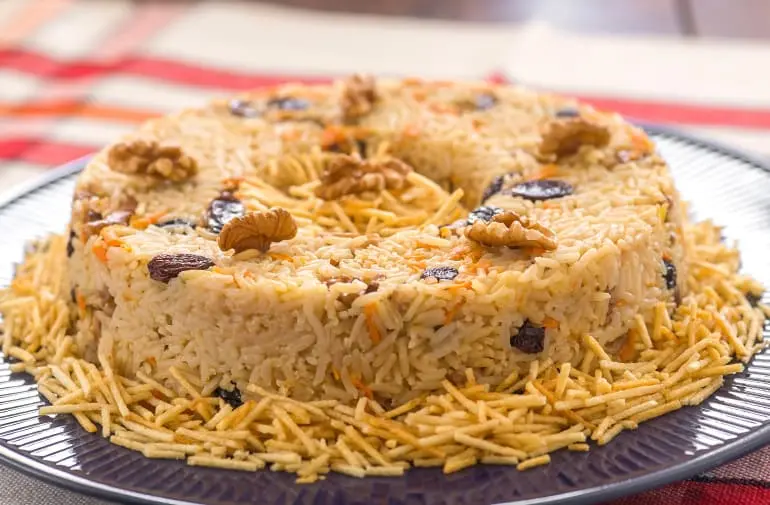 Arroz amarelinho moldado em forma de pudim, com passas e batata palha, decorado com nozes e cenoura, sobre prato preto