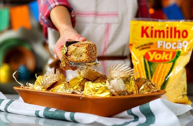 Bolo gelado de milho embalado no papel alumínio, servido em travessa de festa junina, flocão Kimilho Yoki ao fundo