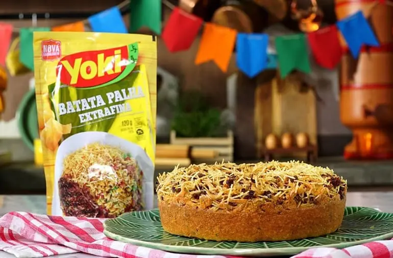 Bolo de milho com carne seca desfiada coberto com batata palha Yoki, servido em prato verde com decoração de festa junina