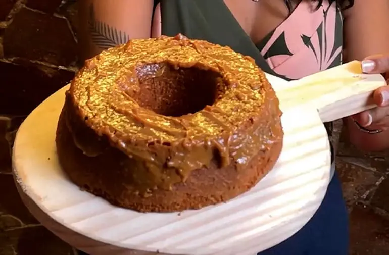 Bolo de tapioca com cobertura cremosa de chocolate, formato redondo com furo no meio, apresentado em tábua de madeira