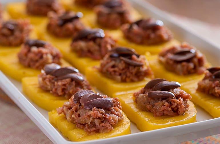 Bandeja com porções de bruschetta de poleta cobertas com calabresa moída e decoradas com azeitona preta