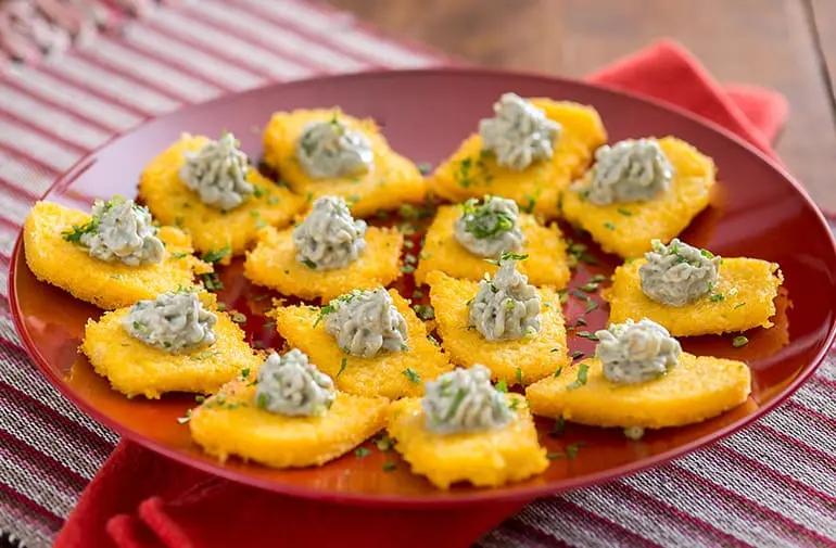 Canapés de polenta frita cobertos por creme de gorgonzola e salsinha, servidos em bandeja vermelha