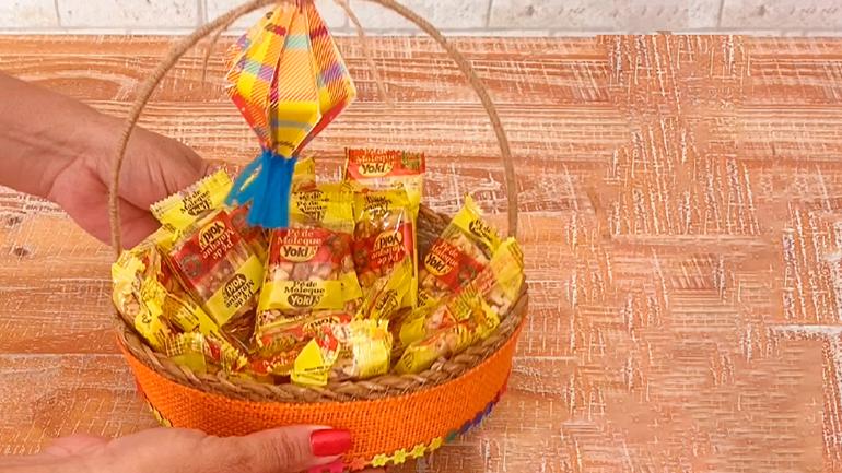 Alguém segura cestinha decorada para festa junina com produtos Yoki e um balão de papel colorido colado na alça