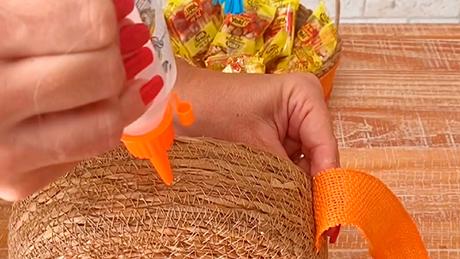 Fita laranja é colada em cesta rústica com cola de silicone, em decoração artesanal de festa junina