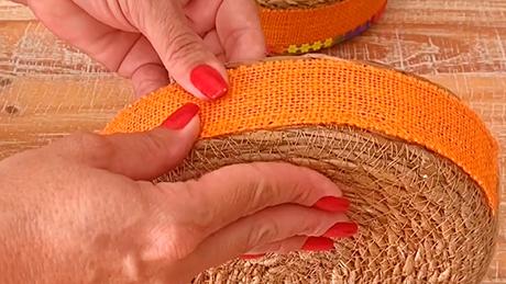 Mãos com unhas pintadas de vermelho apertam fita laranja para fixar a cola em cesta rústica, decoração artesanal de festa junina