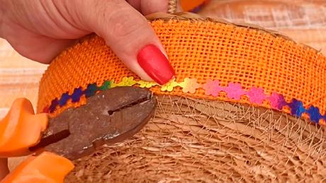 Com ajuda de um alicate, uma pessoa aplica adornos de festa junina sobre uma fita laranja em uma cesta rústica para decoração