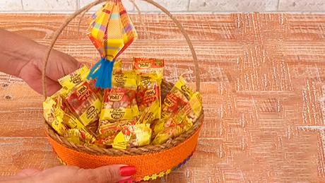 A cesta rústica decorada com balão de papel é colocada sobre a mesa de festa junina, com pipocas Yoki dentro dela