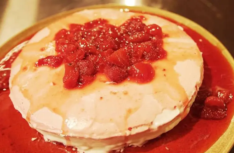 Cheesecake de morango visto de cima, com cobertura de calda de morango e pedaços da fruta, servido em um prato vermelho