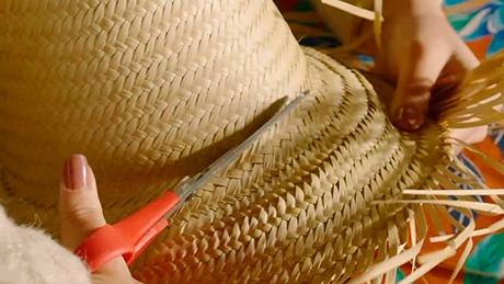 Mãos recortam a aba de um chapéu grande de palha típico de festa junina para fazer decoração artesanal e temática