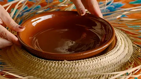 Um prato marrom é colocado no centro de chapéu de palha com topo recortado, formando um sousplat artesanal decorativo