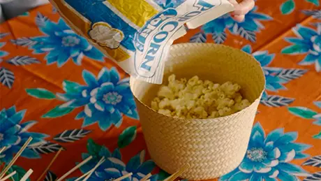 Uma mão despeja o conteúdo de um saco de pipoca de microondas Yoki em cesto feito com o topo de chapéu de palha