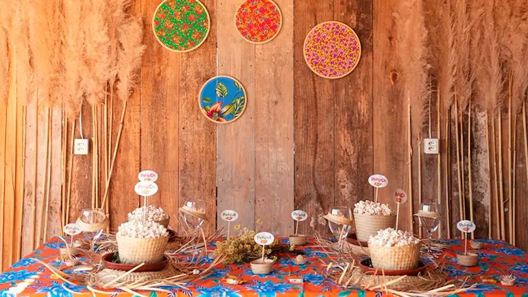 Decoração de festa junina com mesa colorida, comidas típicas em cestos, enfeites e painéis de tecido estampado na parede