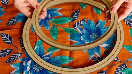 Pessoa posiciona bastidores de tecido sobre toalha floral, preparando decoração de festa junina