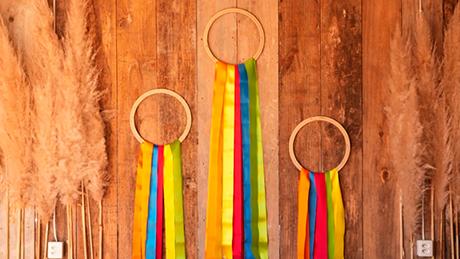 Três argolas com fitas coloridas coladas sobre parede de madeira rústica, em decoração artesanal de festa junina