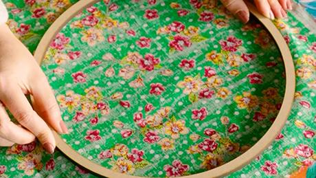 Mãos posicionam bastidor de tecido sobre um pedaço de pano verde com flores, típico de festa junina