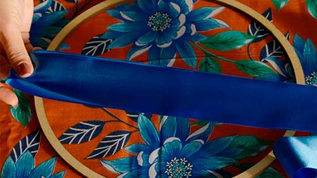 Uma pessoa estica uma fita azul sobre bastidor de tecido pra criar adorno de festa junina