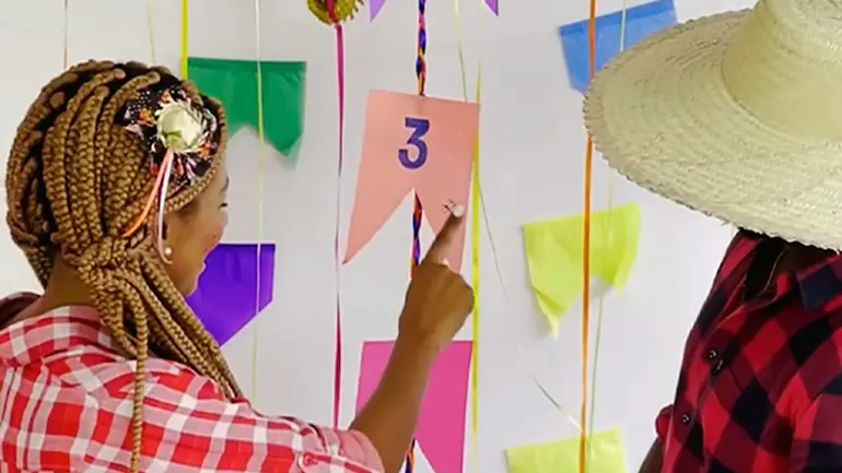 Uma mulher com trajes de festa junina aponta para uma bandeirinha com um número, em brincadeira típica de adivinhação