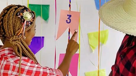 Uma mulher com trajes de festa junina aponta para uma bandeirinha com um número, em brincadeira típica de adivinhação