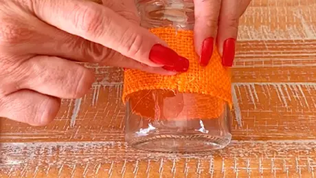 Mãos colam fita laranja sobre potinho de vidro com cola de silicone em atividade de artesanato