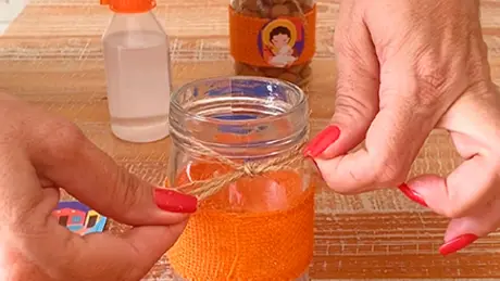 Uma pessoa faz um laço com barbante rústico em volta de potinho de vidro que tem uma fita de tecido laranja colada em torno dele
