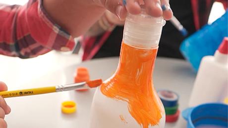 Pessoa com camisa xadrez de festa junina pinta garrafa de plástico com tinta laranja, em mesa de artesanato