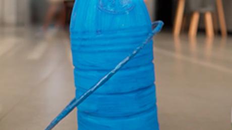 Uma argola é acertada em uma garrafa azul customizada com tema de festa junina, em brincadeira típica