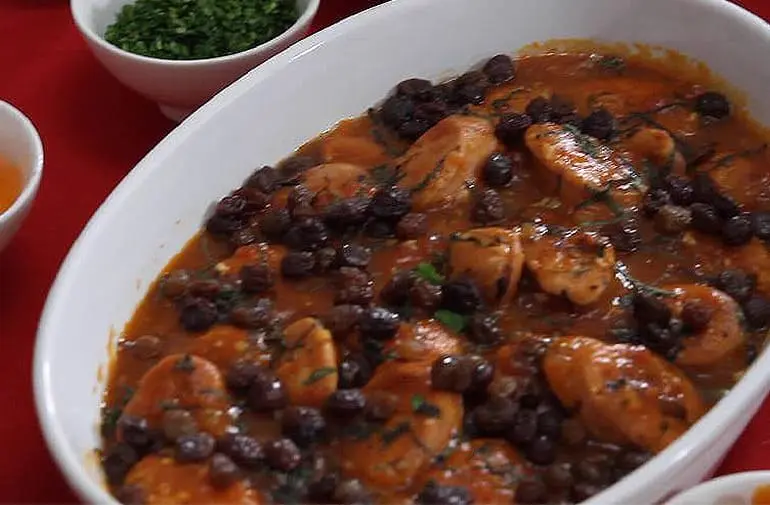 Travessa de lentilha com linguiça de pernil e molho de tomate, próximo a recipiente de temperos em mesa de almoço