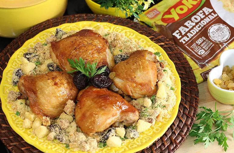 Sobrecoxas de frango assadas e douradas, acompanhadas de farofa de frutas, em mesa de almoço com embalagem de farofa Yoki